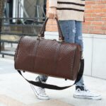 Ein Mann auf der Straße, der eine braune Reisetasche aus Kunstleder an den Henkeln trägt. Darunter hängt ein brauner Schulterriemen. Der Mann steht auf einem Bürgersteig auf der Straße, hinter ihm befindet sich ein Haus mit einem Fenster.