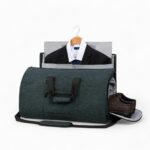 sac de voyage noir chiné type polochon avec anses et bandoulière, avec u ne veste sur ceintre qui sort par le haut ; sur fond blanc.