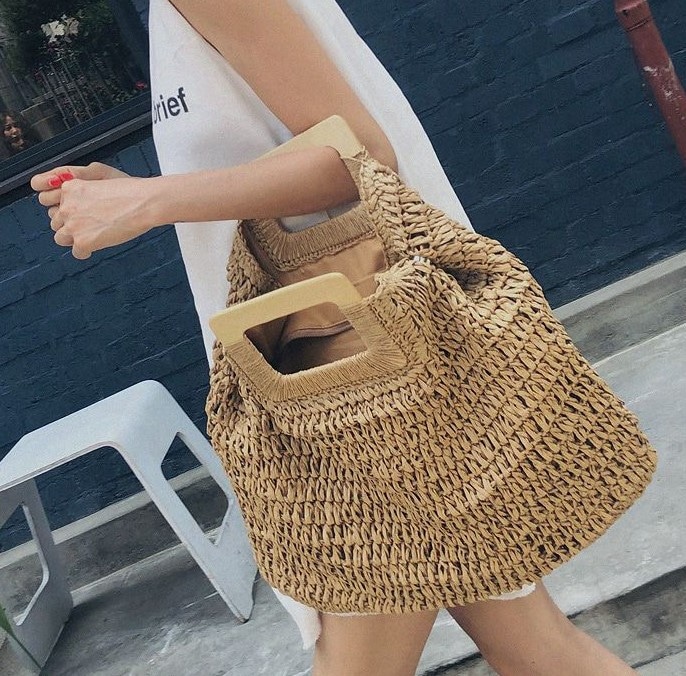 beigefarbene, grobmaschige Strandtasche mit Henkeln aus quadratischem Holz, die mit einem der Henkel über dem linken Unterarm einer jungen Frau getragen wird, von der nur der Oberkörper im Profil zu sehen ist